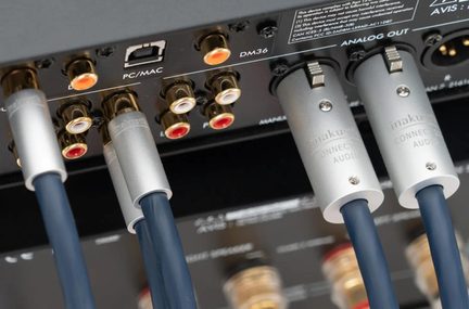 Primo piano leggermente angolato del pannello posteriore di un dispositivo audio, con cavi guainati blu scuro collegati tramite connettori cilindrici in metallo argentato a bloccaggio inseriti in prese RCA placcate oro adornate con anelli rossi e bianchi.