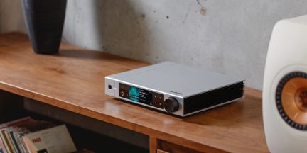 Flacher Hi-Fi-Audioverstärker aus gebürstetem silberfarbenem Aluminium, Dreiviertelansicht von vorne rechts, Front mit 6,35-mm-Kopfhöreranschluss, zentralem Farbdisplay, vier Touch-Tasten und großem schwarzem Lautstärkeregler, rechte Seite mit schwarzem Lüftungsgitter