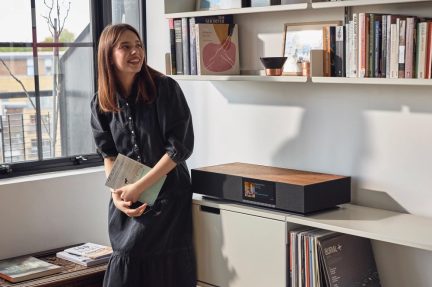 Жінка стоїть, тримаючи книги, полиці з книгами, вікно зліва, меблі з вініловою платівкою, природне світло