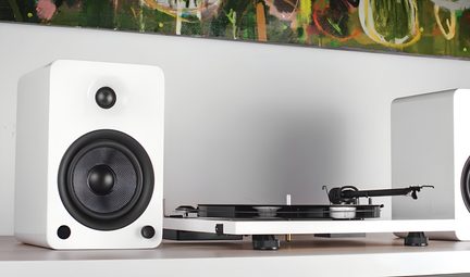 Zestaw audio obejmujący białą, lakierowaną, prostokątną kolumnę głośnikową widzianą od frontu z czarnym wooferem i tweeterem oraz białą gramofon z czarnym talerzem widzianą z boku, ustawione na jasnej półce