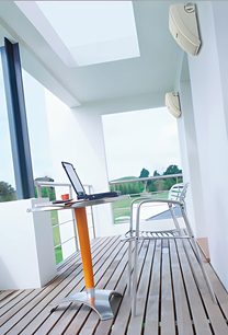 Balkon med hvidt metalskrivebord og -stole, udsigt over have og blå himmel gennem glasparti og hvidt loft