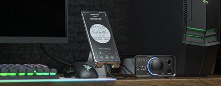 Black smartphone with screen on, placed at an angle on a gray metal stand, surrounded by a black mechanical keyboard with multicolor backlighting, a black mouse lit up in green, and a black audio amplifier with a large blue backlit circular knob, front view on a wooden desk