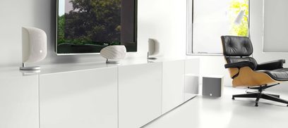 Twee witte compacte Bowers & Wilkins M1 MK2-luidsprekers op een wit meubel, met een zwarte leren fauteuil aan de rechterkant