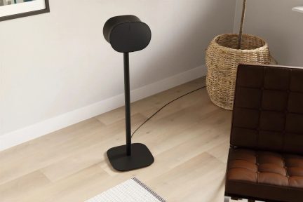 Coluna preta sobre suporte esguio, com frente oval e base quadrada com cantos arredondados, numa sala de estar