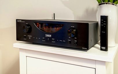 Sort Taga Harmony HTA-2500B hi-fi stereoforstærker, set forfra, stående på et hvidt møbel, med en sort fjernbetjening ved siden af