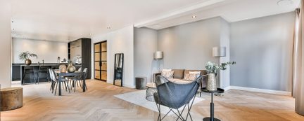 Pohled z úhlu na moderní interiér, světlé dřevěné podlahy v rybinovém vzoru, hladké šedé stěny, obývací kout s pohovkou z béžové látky a černou koženou křeslem motýlek na kovové noze, v pozadí jídelní stůl a černá kuchyňská linka.
