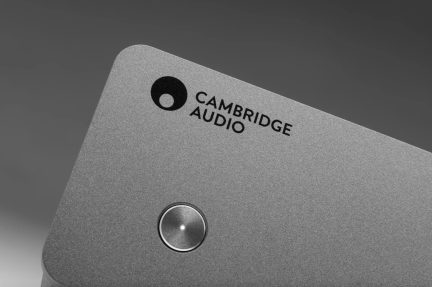Primo piano in leggero controcampo sull’angolo superiore di un case audio grigio argento in alluminio spazzolato, con logo « Cambridge Audio » stampato in nero e pulsante circolare metallizzato con LED centrale
