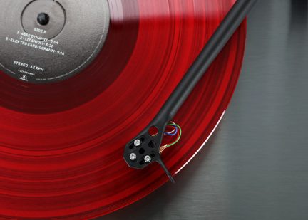 Disco de vinil vermelho translúcido com etiqueta central cinza, braço de leitura preto com cabos coloridos visíveis, superfície metálica