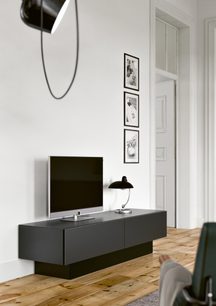 Hængende sort TV-møbel med skuffe, sort skrivebordslampe, tre sort-hvide fotorammer på hvid væg i et moderne interiør