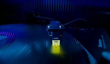 Close-up de uma cápsula fonográfica preta em metal e plástico, ponta amarela com inscrição VNL, sobre um disco de vinil sulcado, iluminação azul