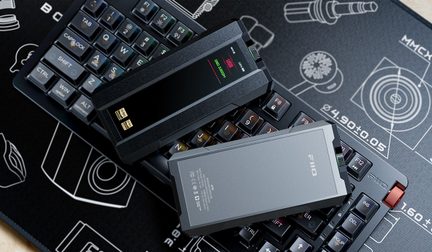 To rektangulære, bærbare lydkabinetter i sort og antracitgrå aluminium med glasfront og farveskærm, gyldne Hi-Res-logoer, FiiO-logo og grøn og rød rillet drejeknap på siden, placeret diagonalt set ovenfra på et mekanisk tastatur