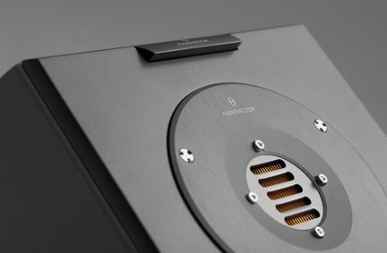 Nærbillede i trekvart fugleperspektiv af en mørkegrå højttaler i børstet metal med et rundt, fastskruet panel med gyldne lamelgitter og Audiovector-logo.