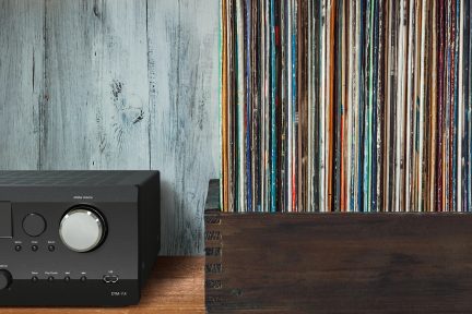 Černý zesilovač s tlačítky a USB portem vlevo, řada vertikálně umístěných vinylových desek v dřevěné krabici vpravo