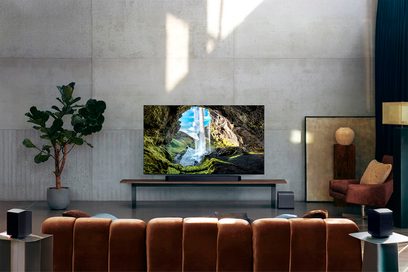 Mattschwarze Soundbar aus Kunststoff mit glatter Frontabdeckung, von vorne unter einem Fernseher auf einem Holzmöbel gesehen, begleitet von zwei schwarzen, würfelförmigen Surround-Lautsprechern und einem passenden Subwoofer