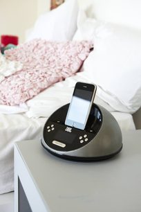 Sort JBL On Time Micro dockingstation, cirkulær form, hvid iPhone placeret ovenpå, på hvidt og lyserødt sengetæppe