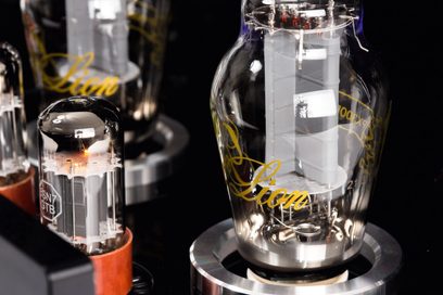 Primo piano obliquo sui tubi elettronici in vetro trasparente, con scritte dorate e bagliore interno arancione, montati su supporti circolari metallici argentati fissati al telaio nero del preamplificatore per cuffie hi-fi Cayin HA-300MK2 nero