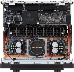Vidinis Marantz AV 20 namų kino stiprintuvo priekio vaizdas: pilkas ir varinis metalinis korpusas, juodos elektroninės plokštės su HEOS moduliu, centre esantis juodas toroidinis transformatorius su auksinėmis raidėmis, matomos vertikalių modulių eilės ir spalvoti laidai.