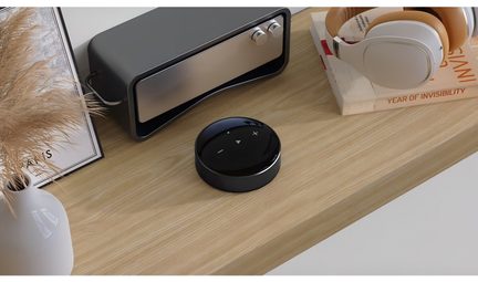 Un disco nero con pulsanti, un dispositivo rettangolare grigio, un casco bianco, un libro e un vaso su una superficie in legno
