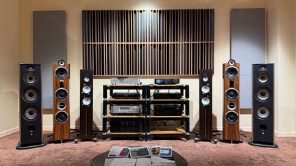 Hi-fi systém s černými sloupovými reproduktory s dřevěným vzhledem, elektronikou na centrálním nábytku a akustickými panely