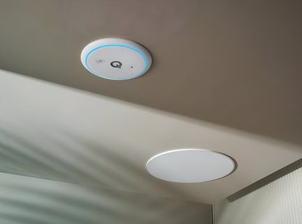 Low-angle view of a beige ceiling featuring a circular white plastic module surrounded by a blue LED halo with a central symbol, next to a built-in speaker with a white metal grille