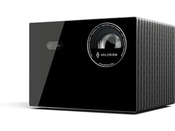 Valerion VisionMaster Max Beamer in Schwarz, glänzende Glasfront mit großem, rundem Objektiv, umgeben von Beschriftungen, und kleinem rechteckigem Sensor, seitliches Gehäuse aus geripptem grauem Metall, Ansicht aus vorderer rechter Dreiviertelperspektive