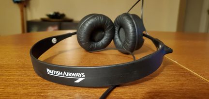 Sort kablet hovedtelefon i plast, ørepuder i sort kunstlæder, hvid « British Airways »-tekst på hovedbøjlen, close-up i trekvart forfra