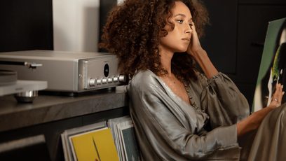 A woman with curly hair gazes pensively downward, seated in front of a silver hi-fi amplifier in a recording studio
