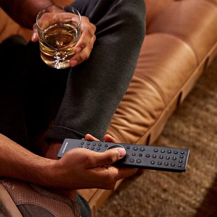 Man's hands holding a glass of whisky and a black remote control on a light brown leather couch