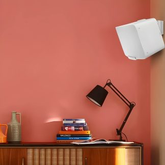 Różowa ściana, biała półka zamocowana, czarna lampa z przegubem, szara karafka, pomarańczowa karafka, stos książek, drewniany mebel