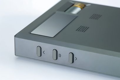 Tæt forfra-venstre perspektivbillede af den bærbare lydkonverter Cayin RU9 i mat grå aluminium, med tre ovale sorte knapper mærket <, pause/afspil og > på siden, en top med et rektangulært glasvindue, en sølvfarvet gittercylinder med gylden ring og to perforerede ventilationsgitter.