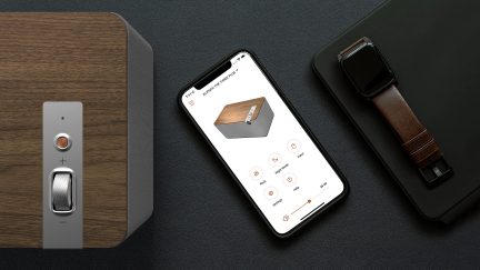 Boxa rectangulară gri, smartphone negru cu interfață de control pe ecran, ceas inteligent maro și negru așezate pe o suprafață neagră