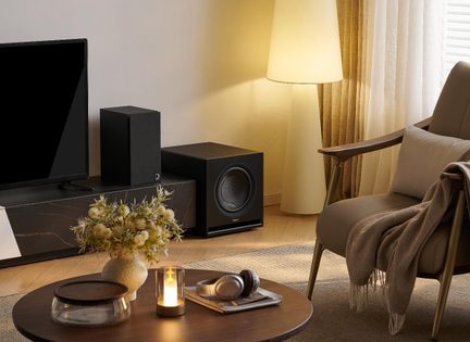 Obývací pokoj, nízký dřevěný stolík s svíčkou, sluchátka a váza, televizor, černá reproduktorová souprava a subwoofer, látková lampa