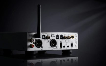 Svetlosivá kovová audio skrinka z pohľadu zozadu, v strede zvislá čierna anténa, dvojité okrúhle XLR výstupy, pozlátené RCA a koaxiálne porty, optické a USB konektory, napájací konektor a logá zhody