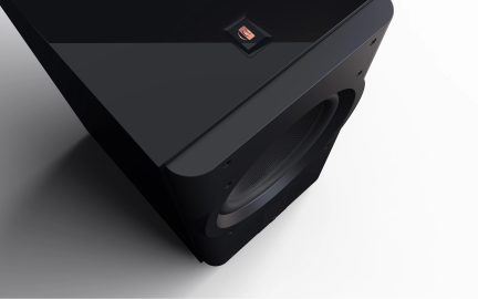 Čierny subwoofer s viditeľnou kruhovou mriežkou, lesklý horný povrch, štvorcové logo v strede, zaoblené rohy.