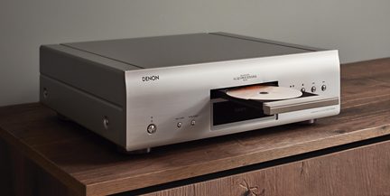 Denon CD player in brushed aluminum silver, three-quarter front view, open tray with disc, display, and front panel buttons