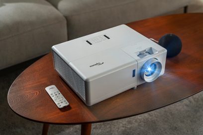 Optoma UHZ50 hvid projektor stillet på et træbord, set fra en trekvart forvisning, med fjernbetjening foran