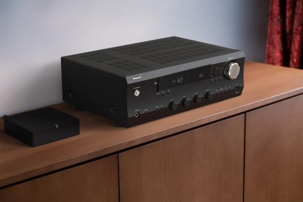 Černý zesilovač s kulatými tlačítky, digitálním displejem, stříbrným knoflíkem, na dřevěném nábytku, pohled ze tří čtvrtin