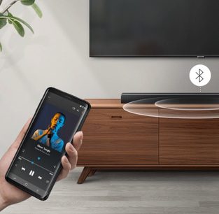 Smartphone, der viser et billede af en mand, der synger i en mikrofon, placeret på et træbord foran en Samsung HW-R530 soundbar