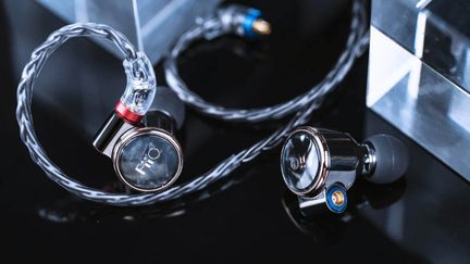 Close-up three-quarter view of a pair of chrome metal in-ear headphones, circular reflective faceplates with white logo, red and blue accents at the connectors, black silicone tips, gray braided cable