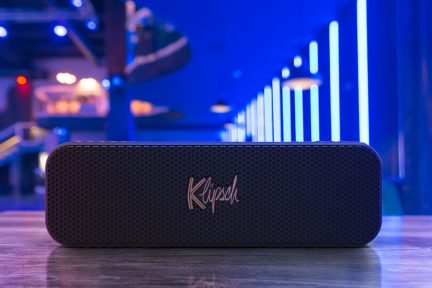Boxa portabilă Bluetooth neagră, dreptunghiulară, cu logo-ul "Klipsch" roz pe un fundal albastru neon, într-un bar sau club de noapte