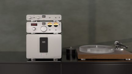 Tre dispositivi audio impilati in metallo argentato con quadranti e pulsanti, giradischi in legno e vetro a destra su una superficie nera