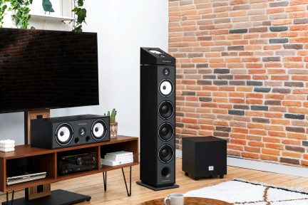 Černý televizor, černý centrální reproduktor, černý sloupový reproduktor, černý subwoofer, dřevěný nábytek, cihlová zeď
