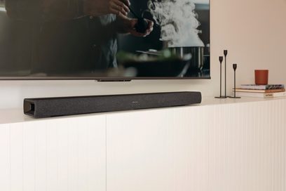 Denon DHT-S217 sort soundbar, set forfra, placeret på et hvidt møbel foran en tændt tv, der viser et sløret billede