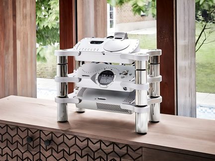 Chasis de audio hi-fi plateado en metal cepillado de tres niveles con columnas cilíndricas cromadas, vista en tres cuartos desde el frente, colocado sobre un mueble de madera