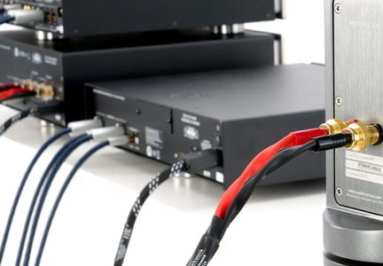 Detailní šikmý záběr na zadní panel z kartáčovaného hliníku s pozlacenými svorkovnicemi propojenými červeným a černým kabelem, před černými audio zařízeními spojenými modrými opletenými šňůrami.