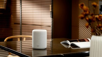Caixa de som conectada WiiM branca em formato compacto, vista de frente sobre mesa escura em um ambiente interno acolhedor