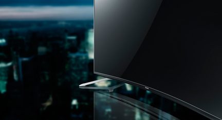 Televisore ultra sottile a schermo piatto visto di profilo, posizionato su un supporto metallico, con una città sfocata sullo sfondo