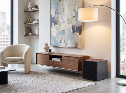 Kompaktní černý subwoofer umístěný na podlaze v moderním obývacím pokoji vedle dřevěného TV stolku