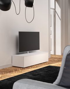 Bílý TV stolek se zásuvkou, pod černou televizí, na parketové podlaze, se dvěma kulatými černými závěsnými svítidly na stropě