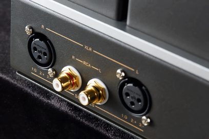 Close-up em ângulo do painel traseiro em metal cinza do Cayin HA-2A, pré-amplificador de fones de ouvido hi-fi, equipado com duas entradas XLR pretas fixadas por parafusos cromados e dois conectores RCA dourados com anéis brancos